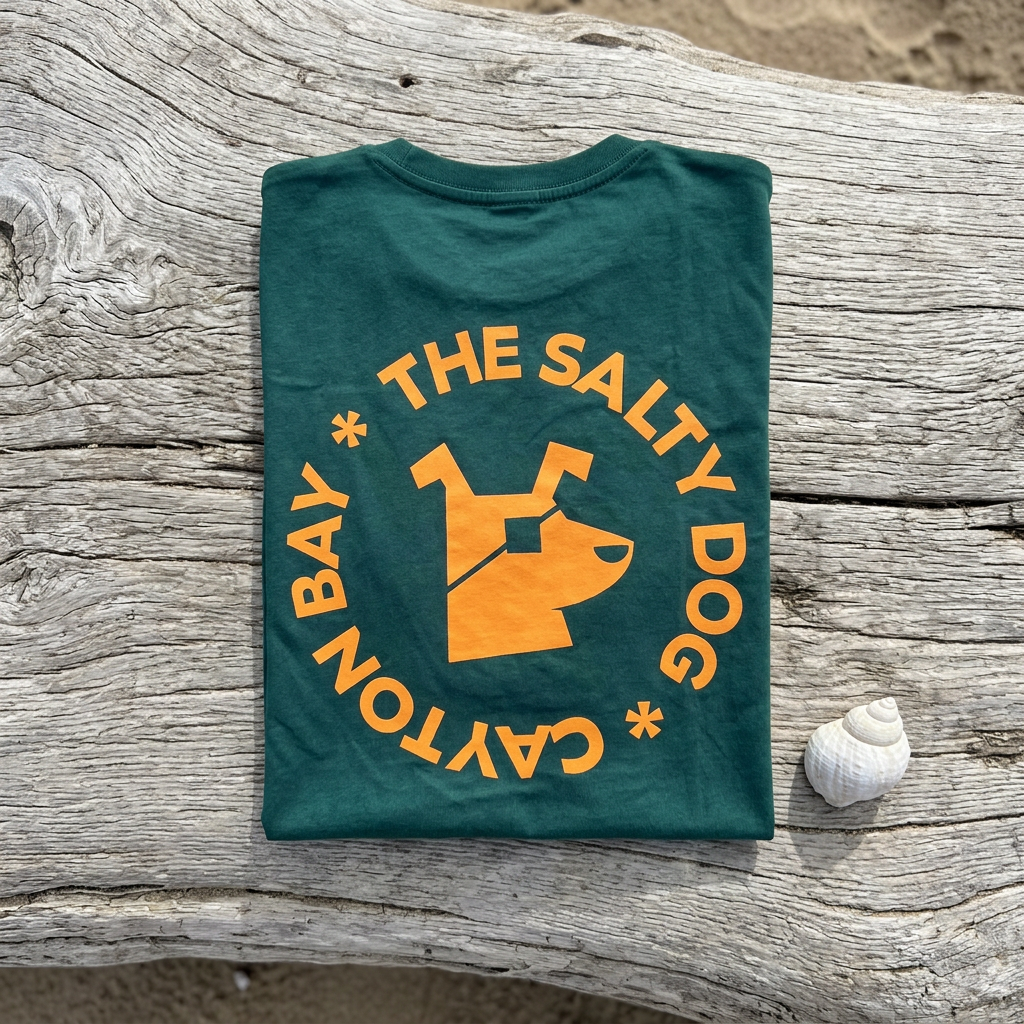Forest green Salty Dog tee folded flat on driftwood with a seashell